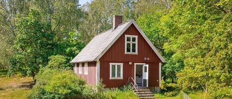 En liten, envåningsstuga med röd fasad, vita detaljer och en skorsten, omgiven av tät grönska.