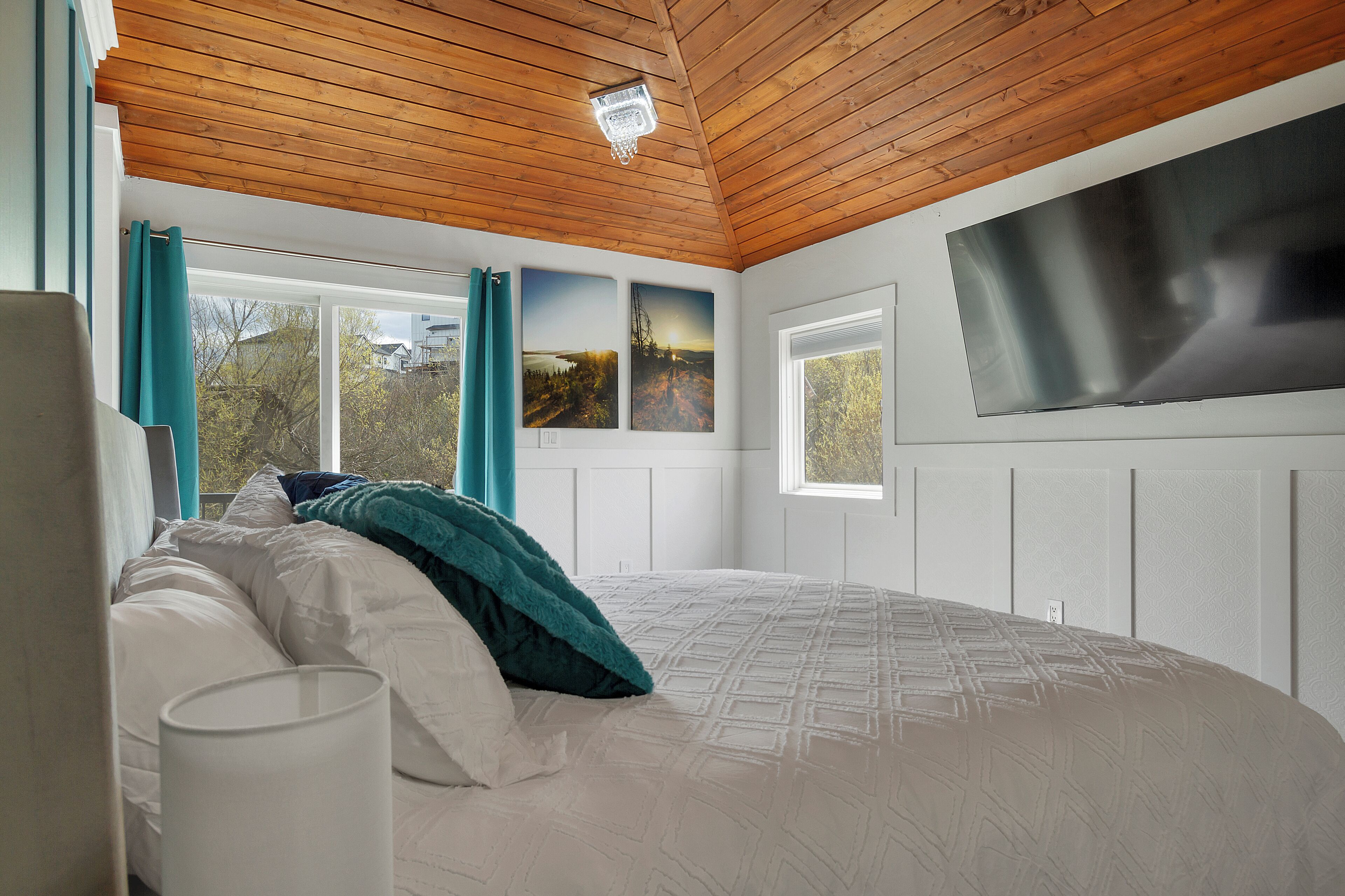Une chambre avec un plafond en bois, un lit recouvert d’une couette blanche et turquoise, deux tableaux représentant des paysages accrochés au mur et une grande fenêtre.