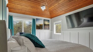 A bedroom with a wooden ceiling, a bed with white and teal bedding, two landscape pictures on the wall, and a large window. - .50 Acre Property - Spacious Living with Wood Accents - Sleeps up to 20 (Vernon)