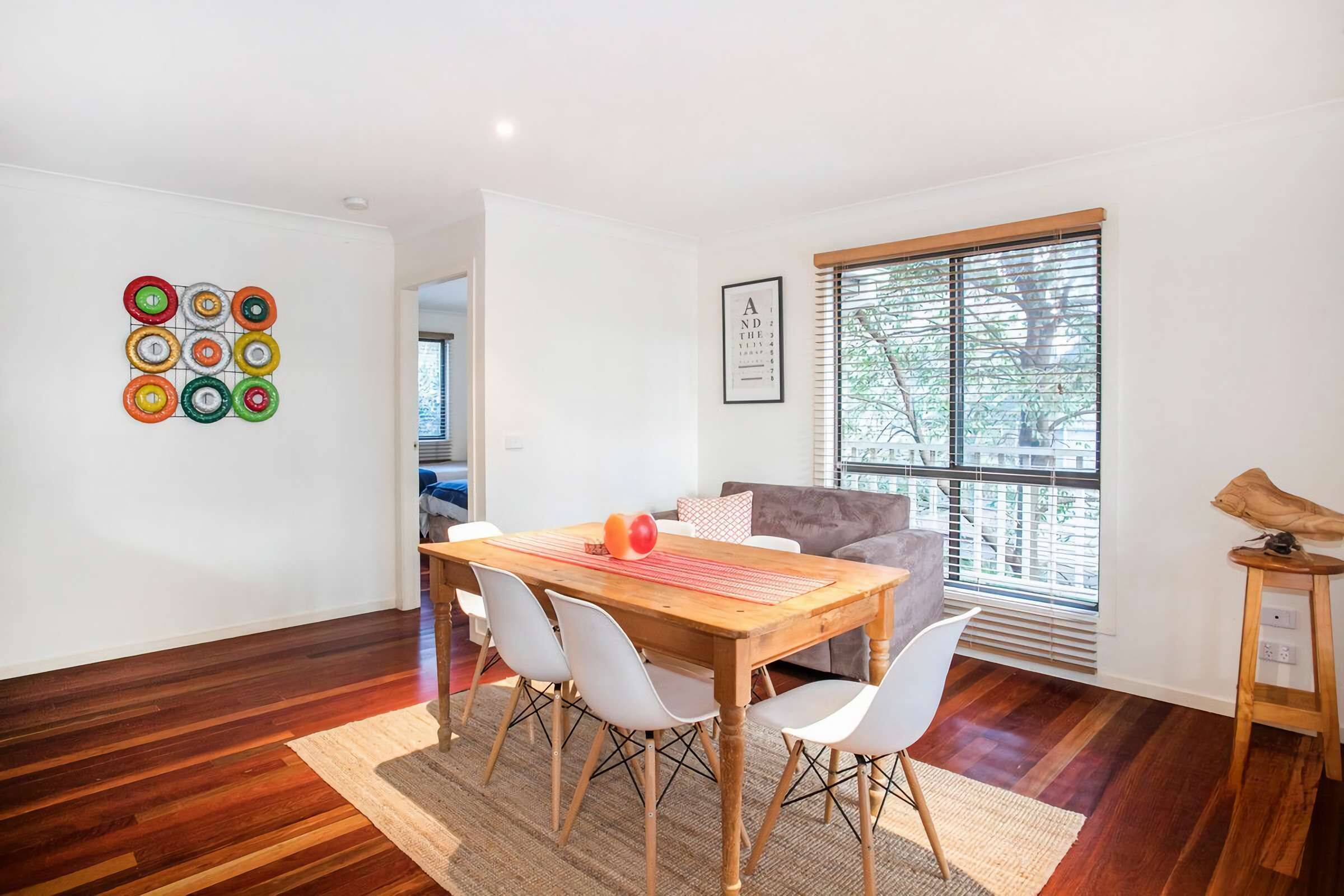Una sala de estar moderna con una mesa de comedor de madera, sillas blancas, un sofá gris y un ventanal con persianas.