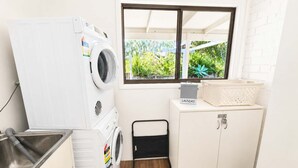 Une petite buanderie comprenant un lave-linge, un étendoir, un petit meuble de rangement et une fenêtre donnant sur un espace verdoyant.