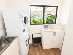 A compact laundry room with a washing machine, a drying rack, a small cabinet, and a window with a view of greenery. - Blue Chill of Mollymook (Mollymook Beach)