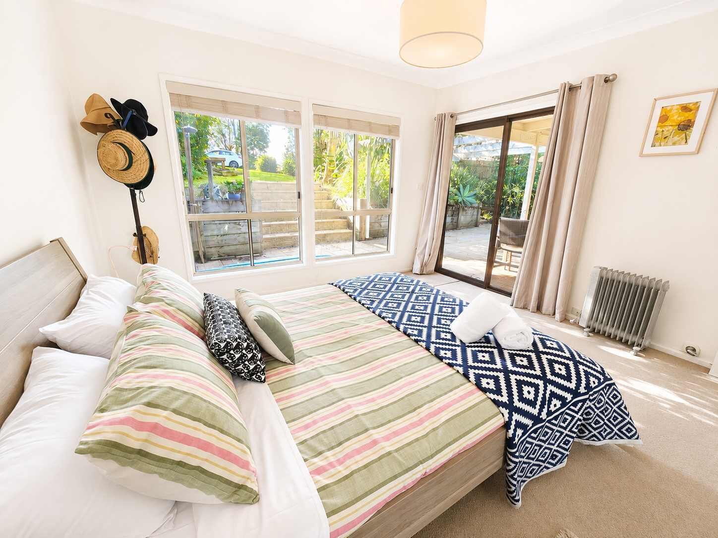 A bedroom with a bed, pillows, a hat on the wall, and a view of the outdoors through large windows.