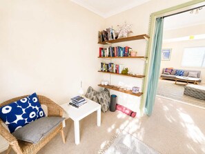 A living room with a wicker chair, a white table, and a bookshelf with books and decorative items. - Blue Chill of Mollymook (Mollymook Beach)