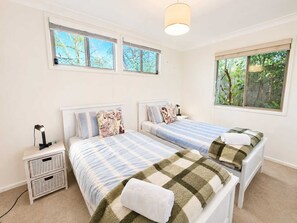 A bedroom with two beds, a nightstand, a lamp, and a window with a view of trees. - Blue Chill of Mollymook (Mollymook Beach)