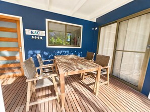 A wooden deck with a table and chairs, a window with a view of greenery, and a sign with numbers. - Blue Chill of Mollymook (Mollymook Beach)