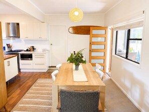 A modern kitchen with white cabinetry, a wooden dining table, and a large window. - Blue Chill of Mollymook (Mollymook Beach)
