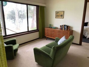 Une pièce avec une grande fenêtre, un fauteuil vert, une commode en bois et un tableau accroché au mur.