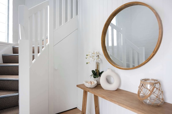 Eine Treppe mit weißen Geländern, ein Holztischchen mit Dekoration und ein runder Spiegel an der Wand.