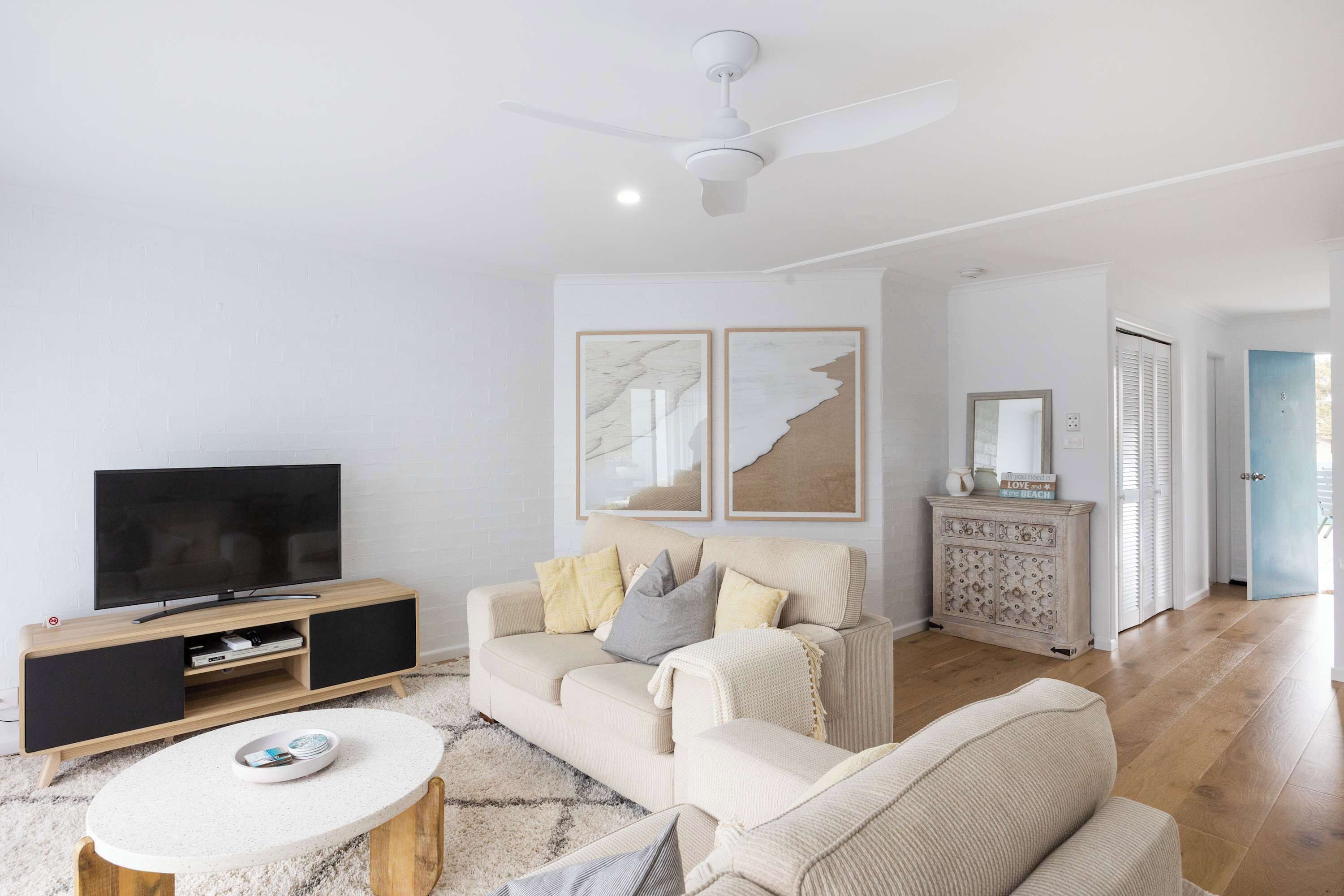 A modern living room with a beige sofa, a flat-screen TV on a wooden stand, a white coffee table, and a ceiling fan.