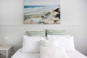 Ein Bett mit weißer Bettwäsche, zwei Kissen, einer gemusterten Tagesdecke und einem Wandbild mit Strandlandschaft.