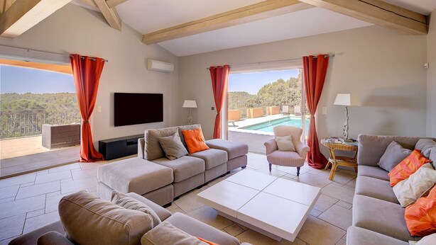 Ein geräumiges Wohnzimmer mit einer großen Couch, einem Couchtisch und einem an der Wand befestigten Fernseher. Zwei Fenster mit Vorhängen und Blick auf einen Swimmingpool und Bäume.