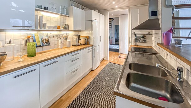 A modern kitchen with white cabinets, a wooden countertop, a stainless steel sink, and a visible hallway leading to another room.