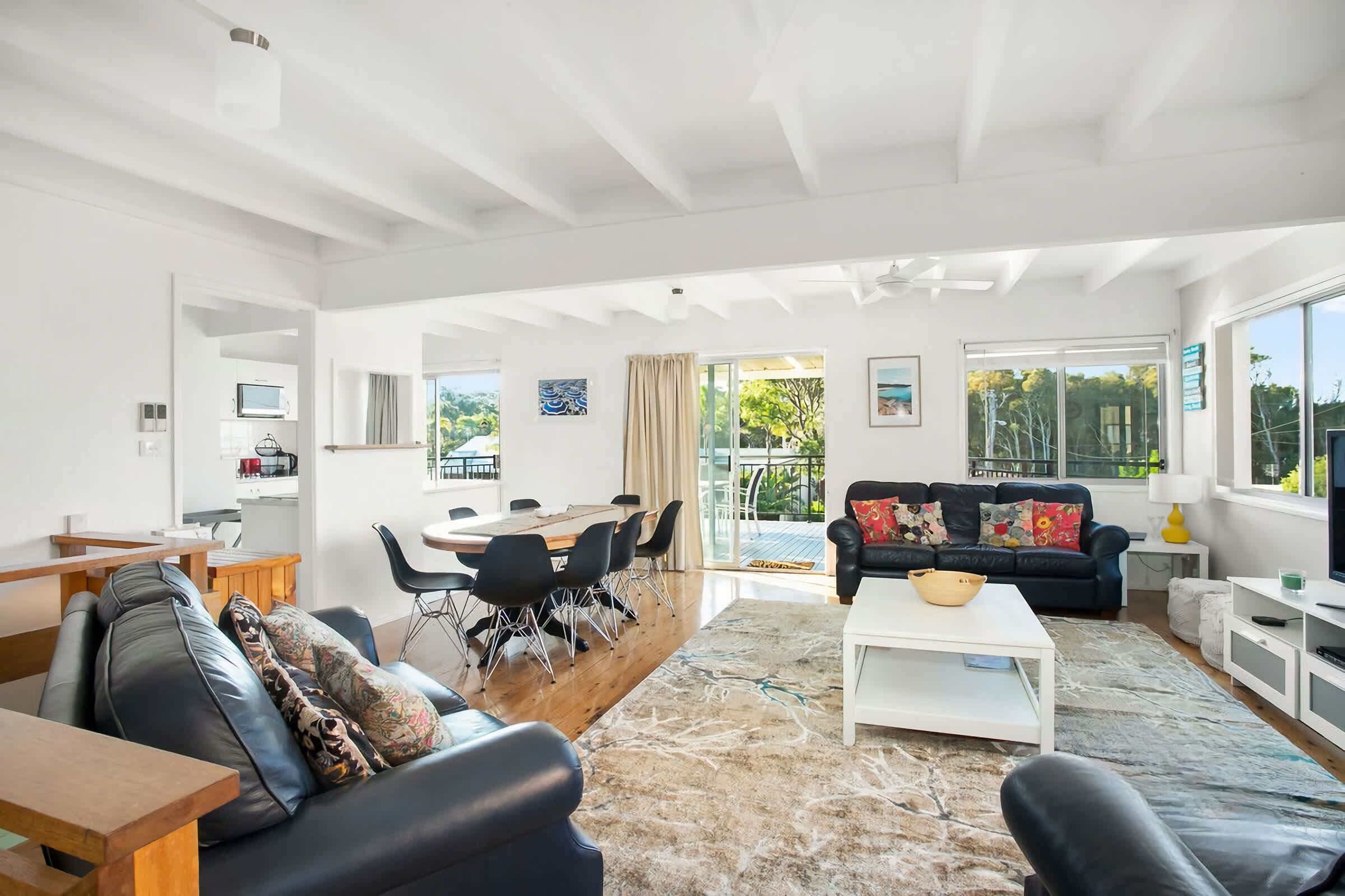 A spacious living area with a black leather sofa, a white coffee table, and a dining table with chairs. There is a large window providing a view of the outdoors.
