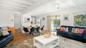 A spacious living area with a large sofa, a coffee table, and a dining table with chairs. A kitchen is visible in the background, and there is a large window providing a view of the outdoors. - MeNo's at Bawley Point (Bawley Point)