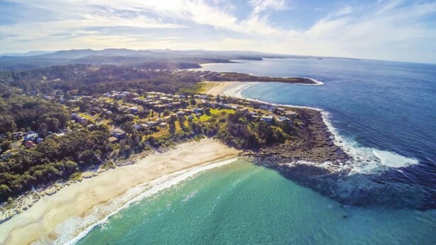 An aerial view of a coastal area with a beach, clear blue waters, and a town with houses and greenery. - MeNo's at Bawley Point (Bawley Point)