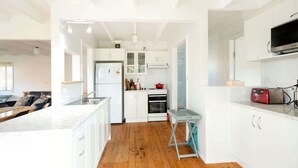 A modern kitchen with white cabinetry, a wooden floor, a built-in oven, and a small dining area with a table and chairs. - MeNo's at Bawley Point (Bawley Point)