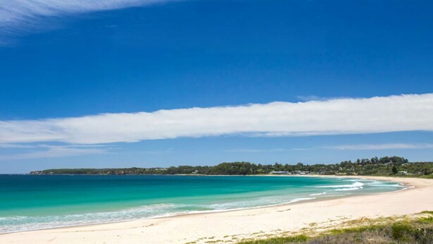 A beach with clear blue water, a sandy shore, and a distant tree-covered landmass under a partly cloudy sky. - Mollymook Beach Cottage (Mollymook Beach)