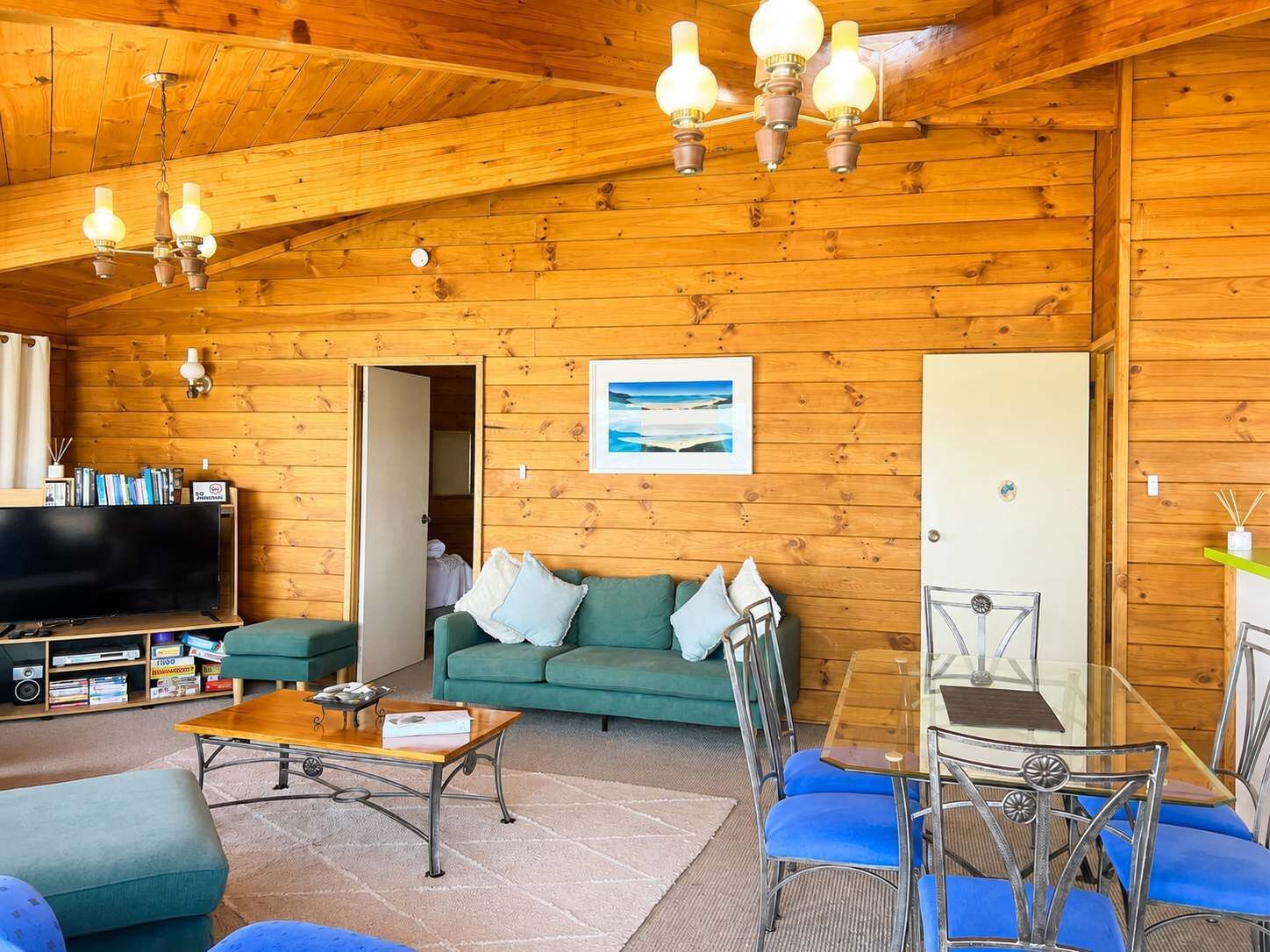 A cozy cabin interior with a wooden ceiling, a green sofa, a wooden coffee table, a glass dining table, and a television.