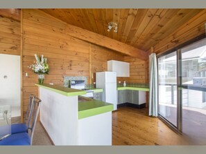A wooden cabin interior with a kitchenette, a dining area, and a large window. - Ocean's Kiss at Mollymook (Mollymook Beach)