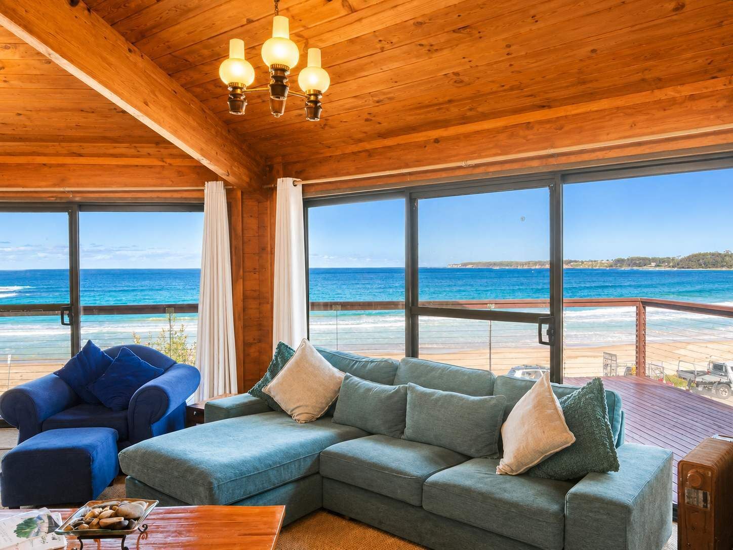 A room with a wooden ceiling, a large window overlooking a beach, a blue armchair, a grey sofa, and a wooden table with a tray of bread.