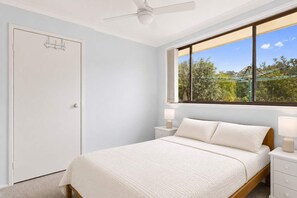 A bedroom with a bed, bedside tables, a ceiling fan, and a window with a view of trees. - Reflections at Lake Conjola (Lake Conjola)