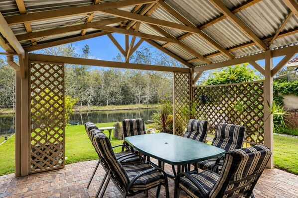 A covered patio with a table and chairs, surrounded by a wooden lattice fence, overlooking a body of water and greenery. - Reflections at Lake Conjola (Lake Conjola)