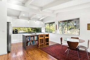 Eine moderne Küche mit weißen Schränken, einer integrierten Bücherwand, einem Essbereich mit rundem Tisch und Stühlen sowie großen Fenstern mit Blick auf Bäume.