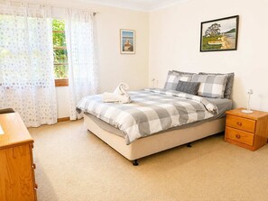 Quarto com cama, mesa de cabeceira, cômoda de madeira, janela com cortinas e duas imagens emolduradas na parede.