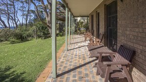 Uma área de estar coberta ao ar livre, com piso de tijolos e bancos de madeira, adjacente a um edifício de tijolos com grandes janelas.