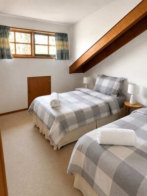 A bedroom with two beds, a window with curtains, a wooden headboard, and a bedside table with a lamp.