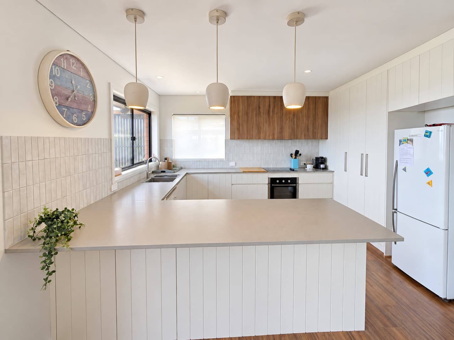 A modern kitchen with a central island, pendant lights, a refrigerator, and a clock on the wall.