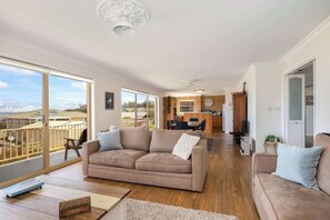 A spacious living room with a large sofa, a coffee table, and a view of the city through large windows. - Rennies Beach Escape "Carinya" (Ulladulla)