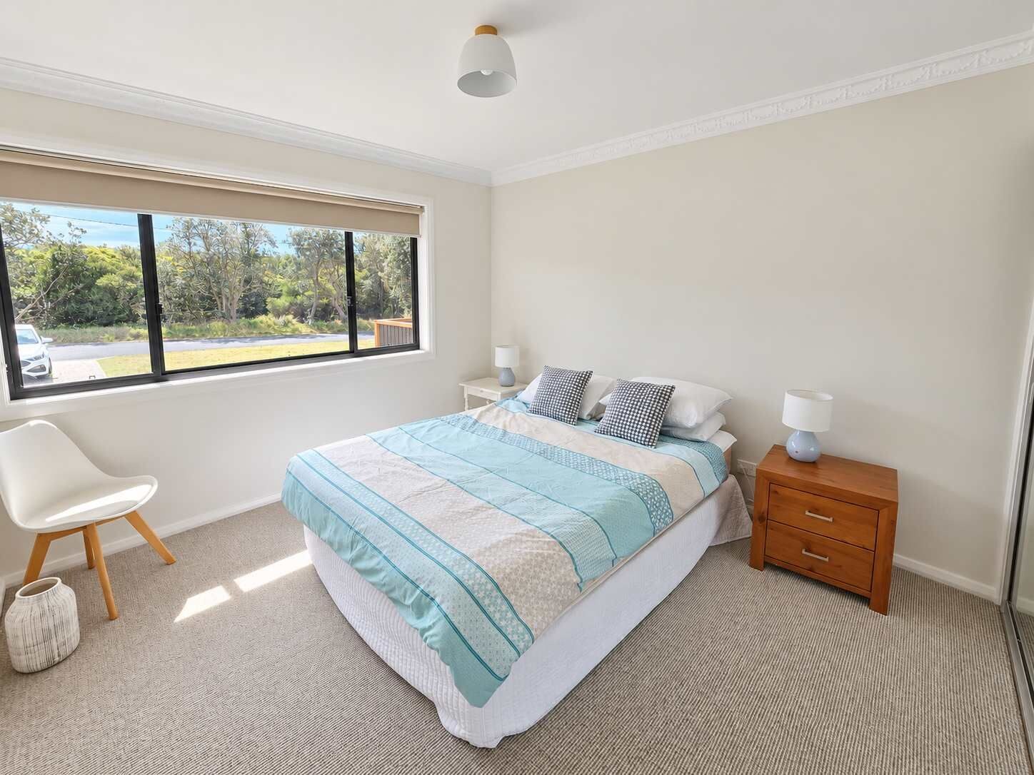 A bedroom with a large bed, two bedside tables, a chair, and a window with a view of greenery.
