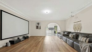 A spacious living room with a large flat-screen TV, wooden flooring, a leather sofa, and a kitchen visible through an archway.