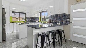 A modern kitchen with white cabinets, a black countertop, and a tiled backsplash. There are three black stools and a window with a view of a building outside.