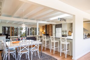 A modern kitchen with white bar stools, a dining table with chairs, and a brick wall. - Narrawallee Beach House (Narrawallee)