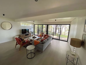 A spacious living room with a large sofa, coffee table, and a television. There is a sliding glass door leading to an outdoor area. - Narrawallee Beach House (Narrawallee)