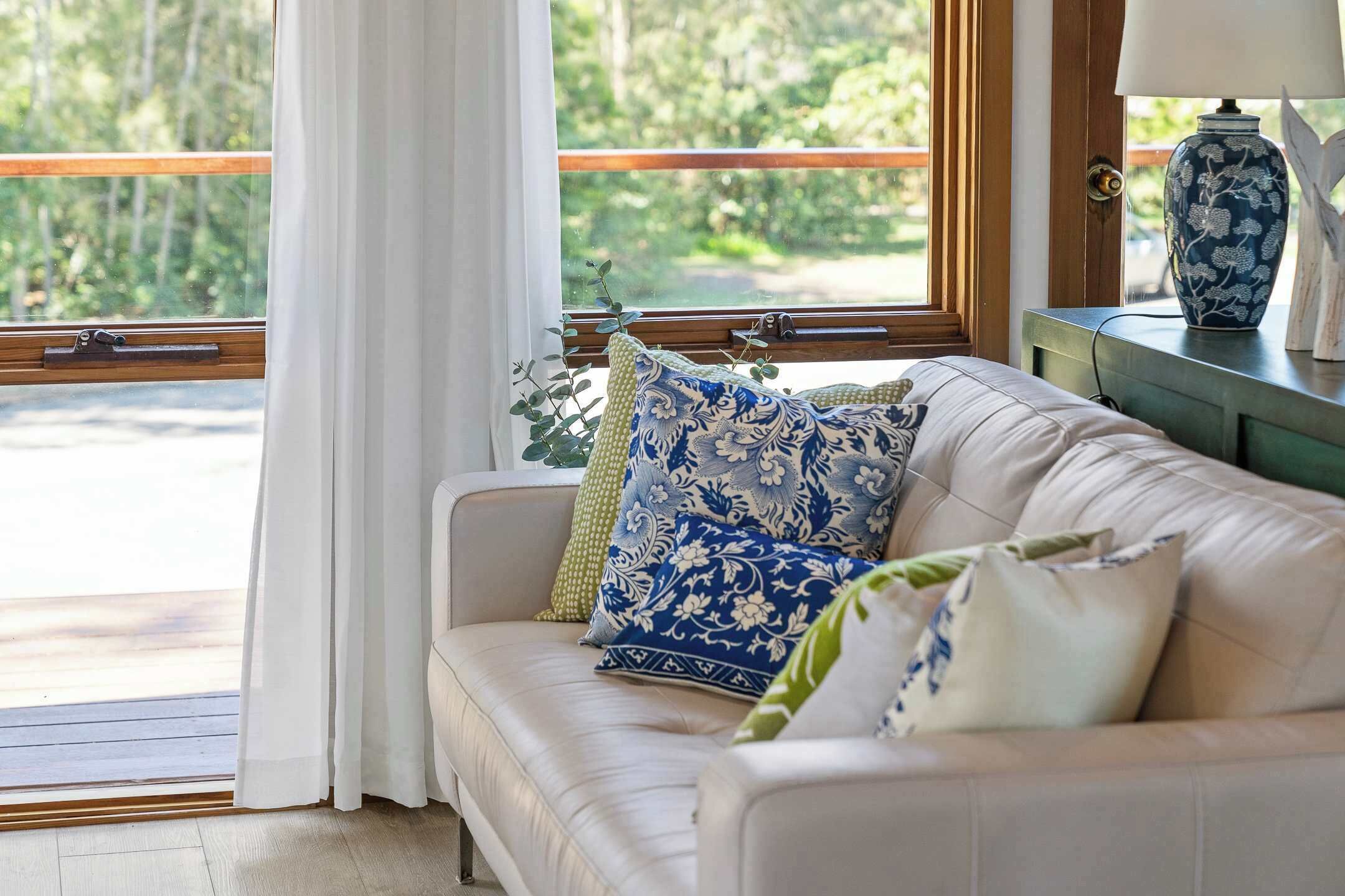 Un sofá de cuero color beige con cojines estampados, una mesita de madera con un jarrón azul, y vista a zonas verdes a través de una ventana con cortinas blancas.