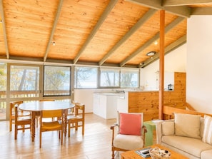 A spacious room with a wooden ceiling, a dining table with chairs, a sofa, and a kitchen area. - Ocean Beach House at Bawley Point (Bawley Point)
