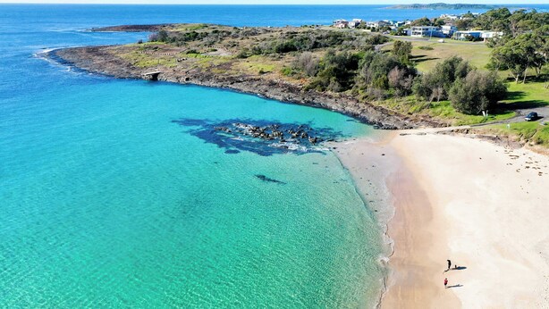 Vista aérea de una zona costera con playa arenosa, aguas turquesas cristalinas y un promontorio rocoso.