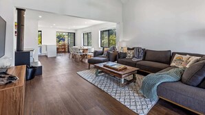 Ein großzügiger Wohnbereich mit dunklem Holzboden, einem großen grauen Sofa, einem Couchtisch und einem Deckenventilator. Im Hintergrund ist eine Küchenzeile mit Esstisch und Stühlen zu sehen.