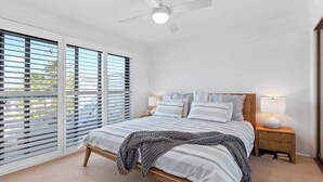 Ein Schlafzimmer mit einem Bett, Nachttischen, einem Deckenventilator und großen Fenstern mit Jalousien.