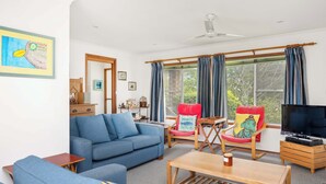 A living room with a blue sofa, a wooden coffee table, and a TV. There are two chairs with cushions, one red and one blue. A ceiling fan is visible, and curtains cover the windows. - Seaview Escape at Gannet (Bawley Point)