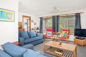 A living room with a blue sofa, a wooden coffee table, a red chair with a cushion, a television, and a window with curtains. - Seaview Escape at Gannet (Bawley Point)