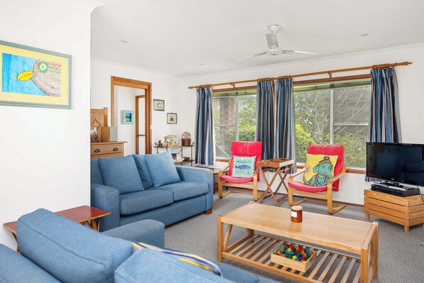 A living room with a blue sofa, a wooden coffee table, and a TV. There are two chairs with cushions, one red and one blue. A ceiling fan is visible, and curtains cover the windows. - Seaview Escape at Gannet (Bawley Point)