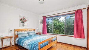 A bedroom with a wooden bed, a bedside table, a lamp, a window with curtains, and a framed picture on the wall. - Seaview Escape at Gannet (Bawley Point)