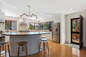 A modern kitchen with a central island, bar stools, a refrigerator, and a view through a sliding door.