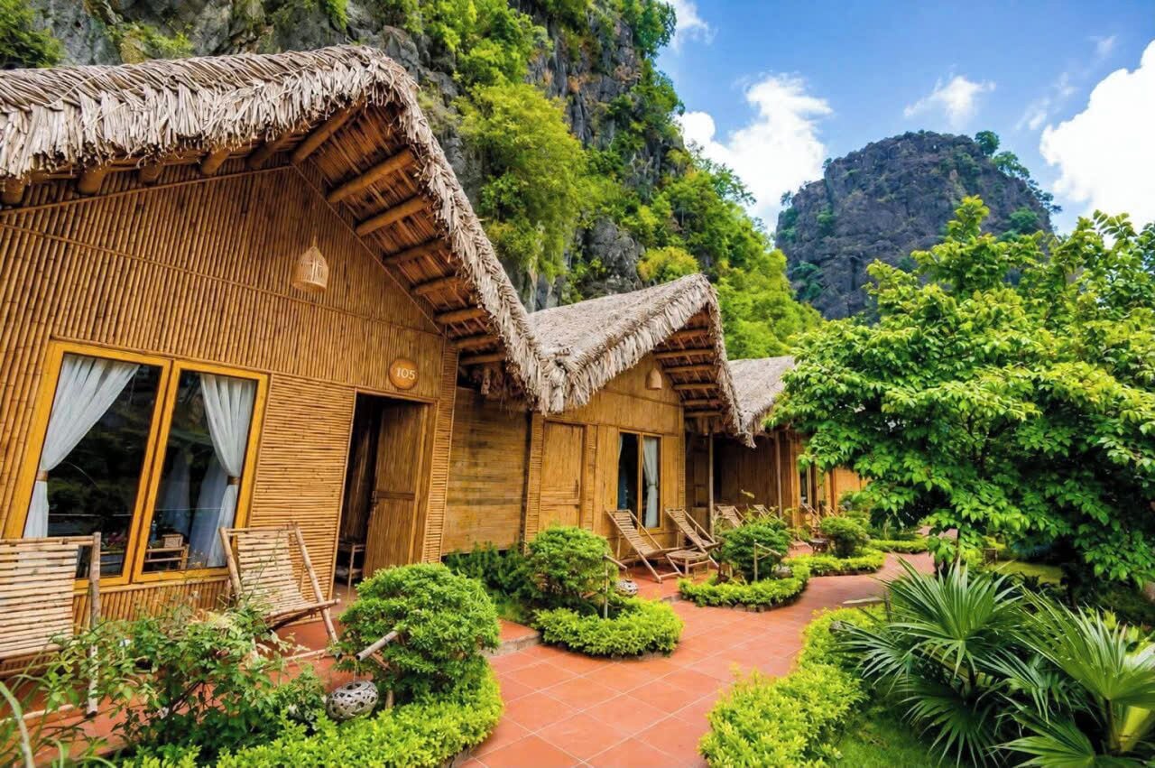 A wooden bungalow with a thatched roof, a veranda with seating, and a lush garden.
