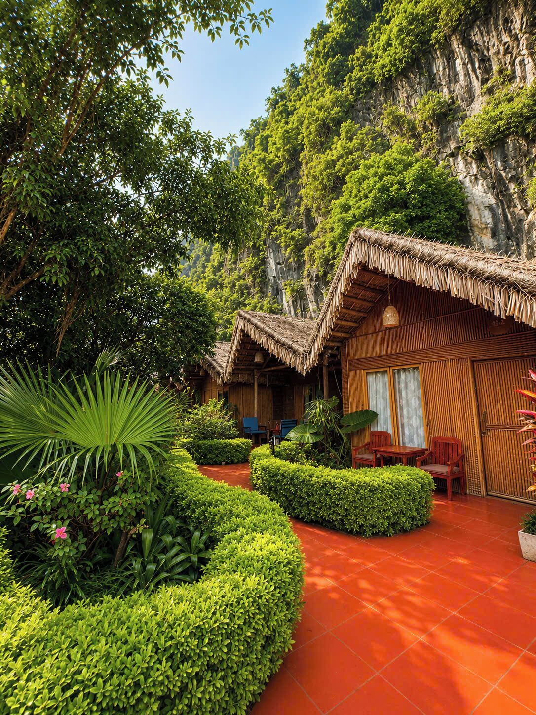 A traditional bamboo hut with a thatched roof, surrounded by lush greenery and a red-tiled pathway.